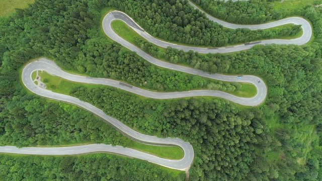 AERIAL, TOP DOWN: Motorcycles and cars driving on zig zag undulating road through lush dense spruce forest on mountain slope. Curvy switchback highway with hairpin turns snaking through the woods