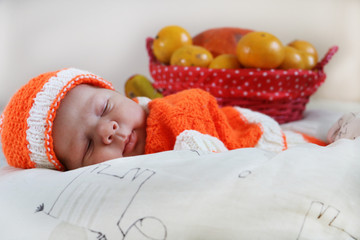 Cute sleeping newborn baby dressed in a knitted orange costume with pumpkin and oranges in basket behind of him. Autumn halloween or harvest concept.