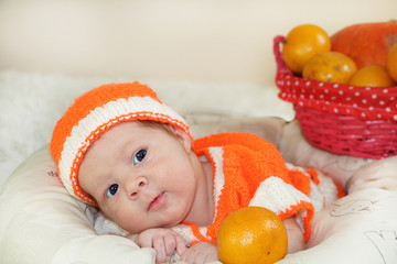 Cute few days newborn baby with funny face dressed in a knitted orange costume with oranges and pumpkin background on white blanket. Baby face expression. 