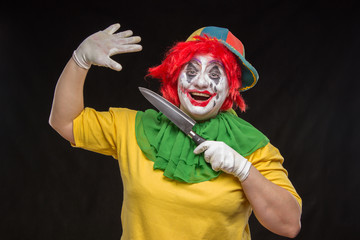 Scary clown with a horrible make-up laughs and with knives in hands on a black background