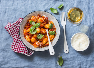 Potato gnocchi in tomato sauce with basil and parmesan and a glass of white wine on blue background, top view. Italian cuisine. Vegetarian food - obrazy, fototapety, plakaty