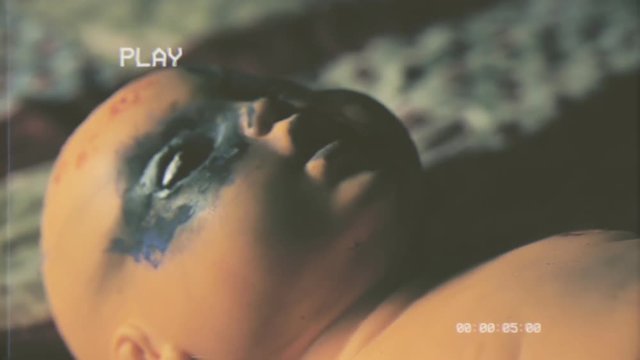 Fake VHS tape: a wicked doll, moving its burnt eyes and mouth, lying on a bed. Handheld travelling shot. Halloween horror themed clip.
