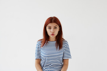 Isolated studio shot of cute teenage girl in striped white and blue t-shirt having startled look, opening mouth and raising eyebrows, thinking what to say. Human facial expressions and emotions