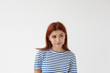 Fototapeta premium Pictures of beautiful young female with red hair pursuing lips and looking sideways with suspicion on her face, raising eyebrow. Suspicious woman feeling distrust toward her unfaithful boyfriend