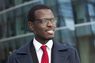 Fototapeta premium Outdoor headshot of young handsome African businessman pictured in urban environment dressed formally in suit, shirt, tie and coat, looking through eyeglasses to street with optimistic smile