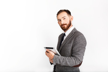 Serious man with red beard poses in grey suit with tablet in his hand