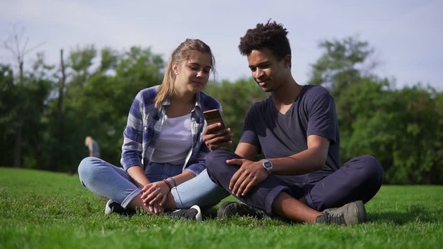 Mixed race couple sitting on the grass and selects pictures in their smartphone. Young people smile and laugh. Cute friends resting in a park, sitting on the grass, enjoying smartphone