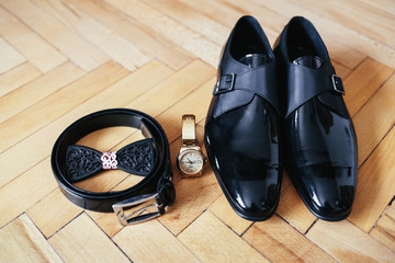 Black leather shoes, golden watch, wooden bow tie and belt stand on the wooden floor