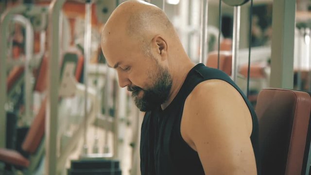 White bald bearded man is doing dips exercise on a machine in gym, orange teal look grade