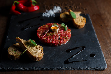 Beef tartar with a quail egg on top, grilled bread, greenery, red chilly pepper, salt and spices, served on a black stone plate.