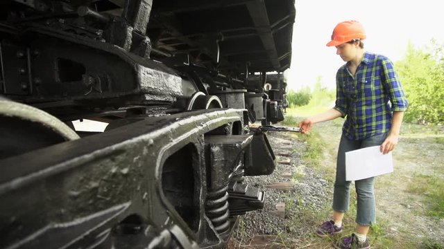 A young brunette woman railway worker in an orange helmet checks the hitch unit of the freight wagon, raises the lid of the axle-box with a plain bearing and compares it with the circuit.