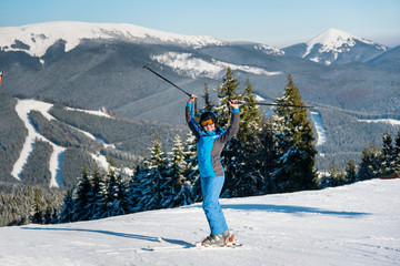 Full length shot of a happy female skier standing on top of the mountains at ski resort, celebrating success holding her equipment up in the air, smiling joyfully happiness enjoyment sports concept