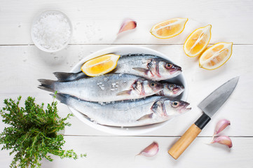 Raw fresh fish on the wooden table in the white plate and kitchen knife, greens, cut lemons