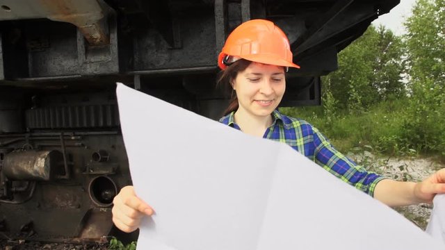 A young woman worker in orange helmet stands near a mining excavator and looks at the project then folds it and smiles.