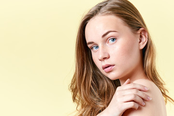 Naklejka premium Pretty redhead teenager girl with blue eyes and freckles, with naked shoulders, looking at camera. Model with light nude make-up, yellow background. Beauty portrait with copy space.