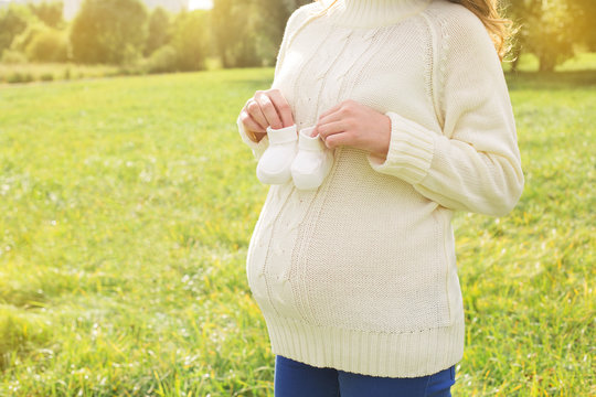 Pregnant woman walking in park sunny summer outdoor mother touching tummy holding little baby shoes
