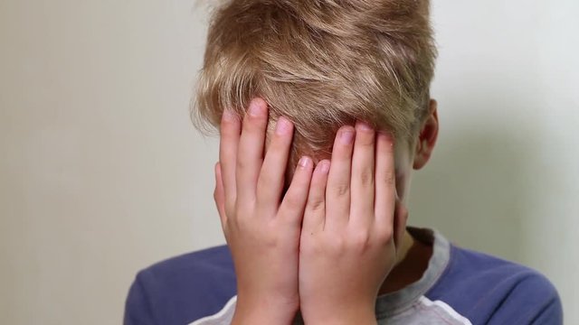 Closeup portrait of face of unhappy white kid covered his face with both hands. Sad lonely little caucasian kid isolated at white wall background in front of camera. Real time full hd video footage.