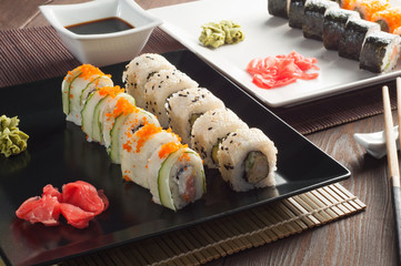 Sushi rolls on a black and white plate, on a wooden mat with chopsticks and soy sauce