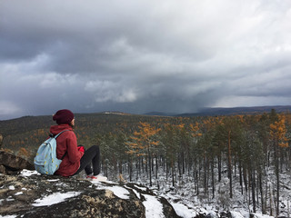 A young athletic girl in warm bright clothes and a hat sits on top of a mountain and looks at a fabulous beautiful bright autumn golden forest in the snow and a misty stormy sky.