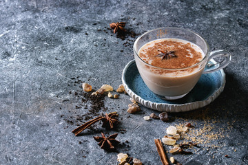 Glass cup of traditional indian masala chai tea on blue saucer with ingredients above. Cinnamon, cardamom, anise, sugar, black tea over dark texture background with space