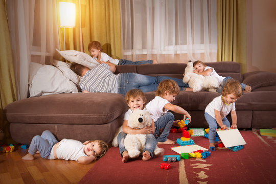 Tired The Father Lies On A Sofa In The Room. Child Scattered Toys. Mess In The House. A Lot Of Children. The Little Children Play In The Room.