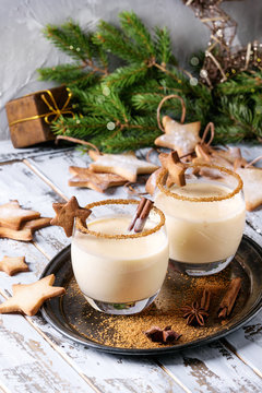 Eggnog Christmas milk cocktail with cinnamon, served in two glasses on vintage tray with shortbread star shape sugar cookies, decor toys, fir branch over white wooden plank table.
