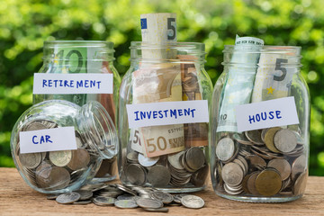 group of glass jar bottles with full of coins and banknotes labeled as investment, house, car and retirement as savings or investment concept