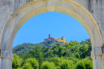 Fototapeta na wymiar Close up of Pena National Palace in the blue sky surrounded by green hills, seen from the arch of Seteais Palace. Sintra historic center, Lisbon District, Portugal.