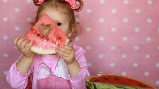 Funny cheerful happy little girl holding a piece of watermelon with a pattern in the form of a heart. Happy child close-up of playing and looking at camera. St. Valentine's Day