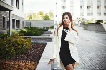 Beautiful young fashionable woman in a black short dress and white cardigan posing in the city. Fashion photo