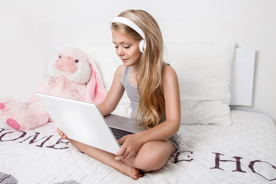 Gracious young beautiful blonde girl, little princess with long hair and blue eyes, the child lies in bed, listening to music, playing the game on a laptop computer and smiles joyfully