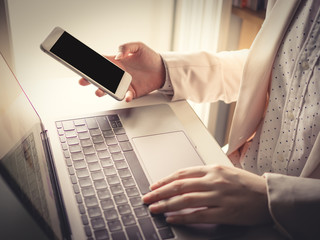 close up smart phone on right hand of business woman(30s to 40s) with pink or pastel suits working with her computer laptop with soft focus left hand and foreground keyboard
