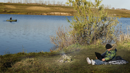 Fototapeta na wymiar the boy is sitting on the shore of a reservoir with a laptop in his hands in the autumn afternoon