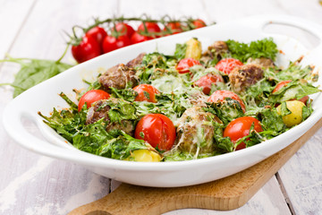 Meatballs and spinach oven dish with cherry tomatoes, potatoes and herbal cheese and cream sauce. One pot meal on White wood background.