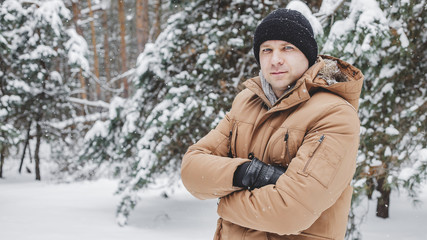 Young man in coat posing with hands crossed and smiling at camera on background of snowy forest.