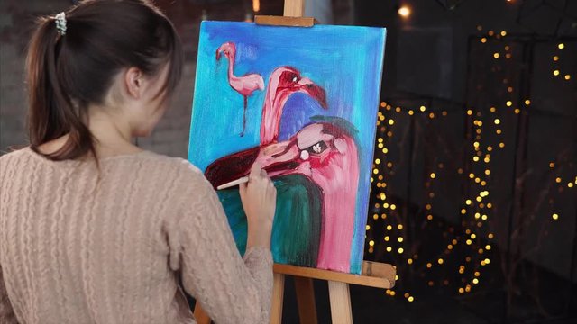 One skilful girl is drawing three cute flamingos on an easel. She is done with a backgroung and now working on a details.