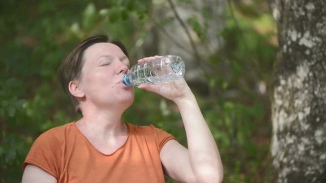 Woman drinking water outdoors. Thirsty adult caucasian female refreshing after long walk through park on sunny summer day.