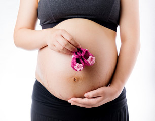 Happy pregnant woman take pink wool baby shoes in hand and touch belly, isolated on white background