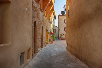 Obraz premium Beautiful narrow street with sunlight and flowers in the small magical and old village of Pienza, Val D'Orcia Tuscany, Italy.