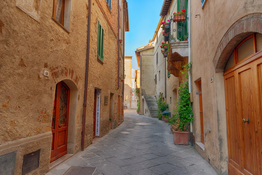 Fototapeta Beautiful narrow street with sunlight and flowers in the small magical and old village of Pienza, Val D'Orcia Tuscany, Italy.