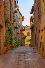 Beautiful narrow street with sunlight and flowers in the small magical and old village of Pienza, Val D'Orcia Tuscany, Italy.