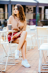 Vertical full height photo of teenager hipster fashionable woman spending time alone sitting in street cafe in early morning. Weekend, travel and leisure concept.
