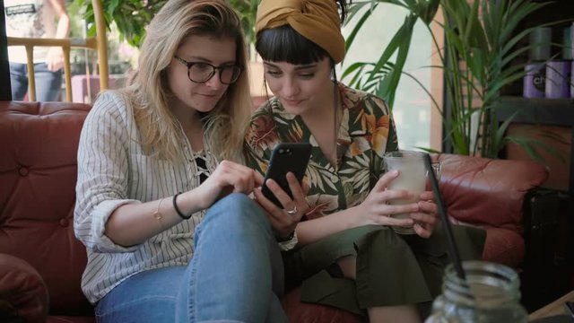 Soft focus hand held shot of two pretty woman, brunette and blonde, young hipster millennials sit in cool cafe coffee house, scroll through dating applications and discuss photos and chat