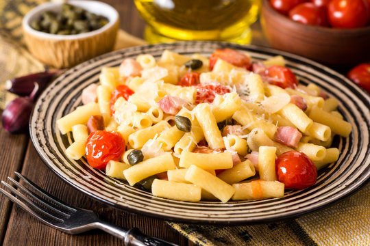 Pasta with bacon, garlic, tomatoes, capers and parmesan cheese in plate on dark wooden background.
