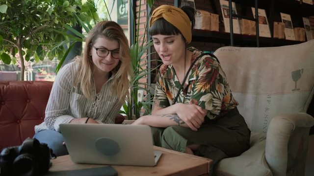 Two beautiful women, brunette and blonde, digital natives, millennial hipsters use video chat application on laptop inside busy trendy cafe, talk and have conversation with family or friends