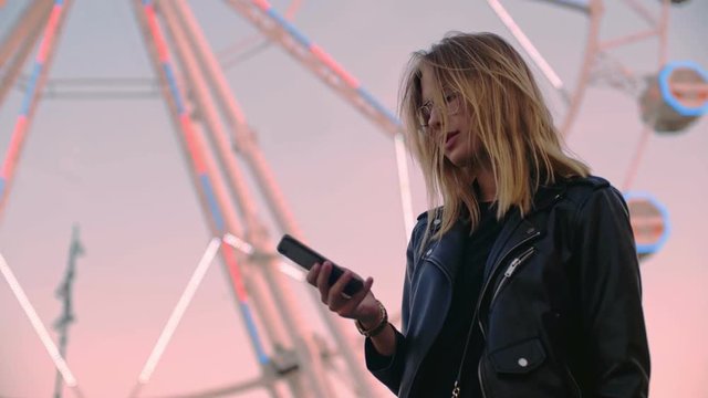Serious and concentrated attractive pretty cute young beautiful blonde woman or teenager in leather jacket stands in front of ferris wheel ride carnival attraction, chats texts on phone or social