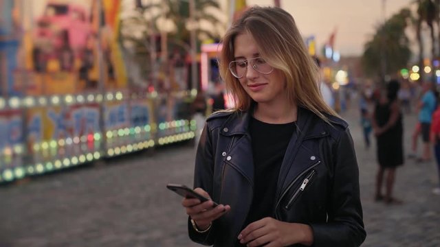 Pretty beautiful attractive woman in hipster millennial glasses uses smartphone to chat and meet up with friends on messenger application, looks around and smiles into camera