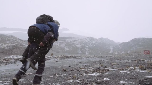 Young man carrying friend on shoulders at mountain in a snow storm. Slow motion