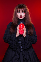 Beautiful woman, a witch, a vampire holds a heart, on a red background. American, dress for a costume party Halloween. Bright makeup