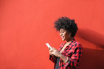 Cheerful girl using her smart phone, standing in front a red wall - obrazy, fototapety, plakaty